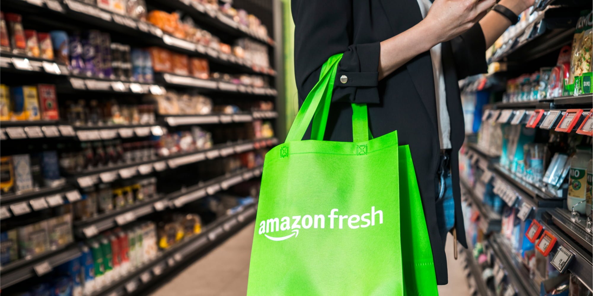 A person holds a lime green Amazon Fresh reusable bag in a grocery store aisle. 