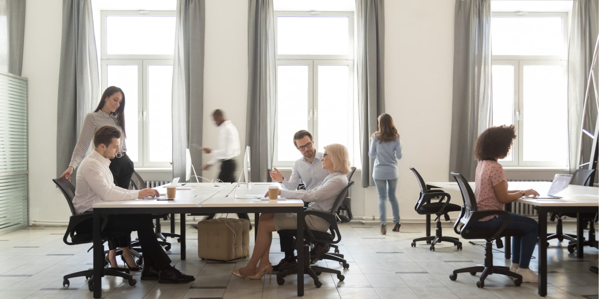 Busy company department members employees working together sitting at shared desks use computers walking in modern coworking space room interior daylight through windows